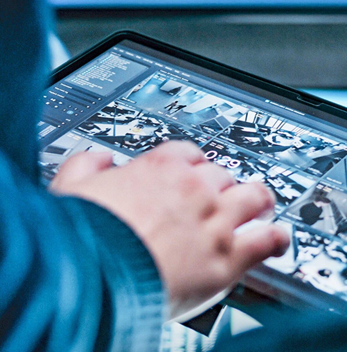 A person interacts with a touchscreen tablet displaying multiple security camera feeds. The background shows a blurred control room setting with monitors.