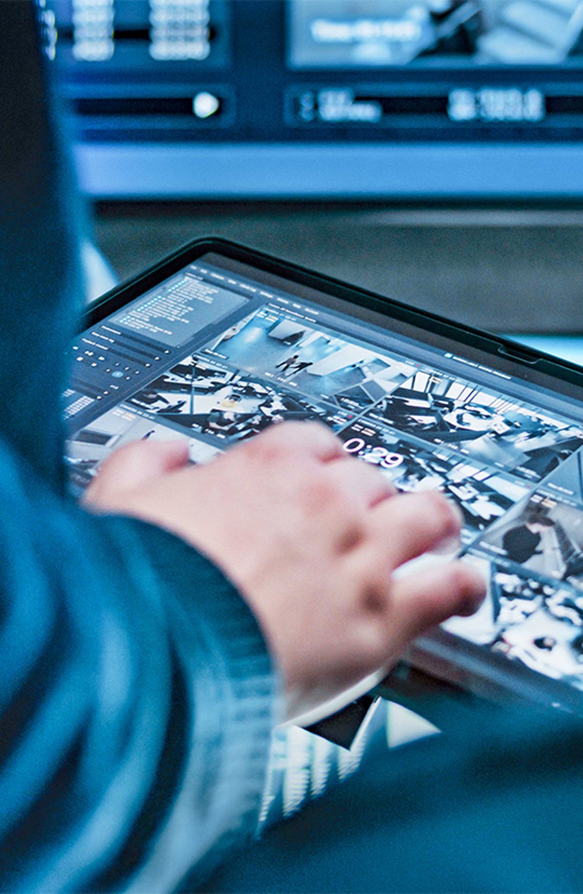 A person interacts with a touchscreen tablet displaying multiple security camera feeds. The background shows a blurred control room setting with monitors.