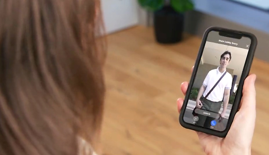 A person holds a smartphone displaying a video call with another person wearing a white shirt and a crossbody bag. The background shows a wooden floor and a potted plant.