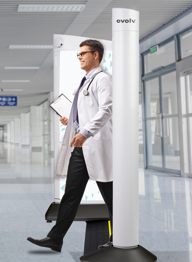 A doctor in a white coat and stethoscope walks through a security scanner in a hospital hallway, holding a clipboard. The scanner is labeled "evolv." The hallway is bright with glass doors and a blue sign in the background.