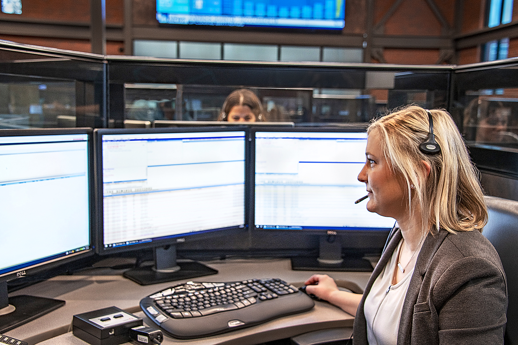 A woman wearing a headset sits at a desk with multiple computer monitors displaying data. She appears focused and engaged in her work. The background shows an office environment with another person visible behind glass panels.