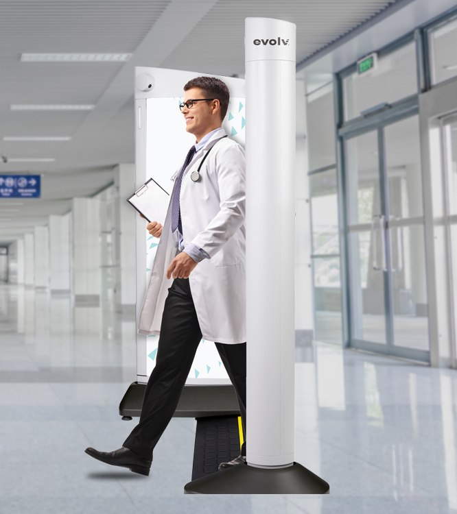 A doctor in a white coat and glasses walks through a security scanning device in a hospital corridor. He holds a clipboard, and the background shows a modern, well-lit hallway with glass doors and signage.