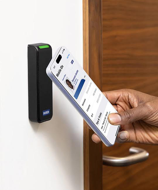 A person holds a smartphone displaying an ID card app near a wall-mounted access control reader. The reader has a green light, indicating access granted. A wooden door with a silver handle is partially visible in the background.