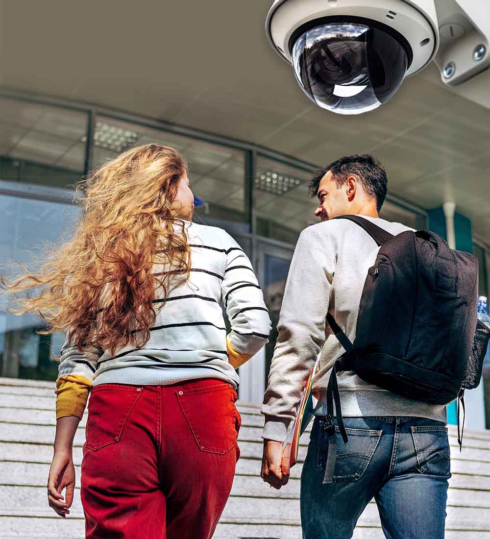 A woman and a man, both casually dressed, walk up steps towards a building entrance. The woman has long hair and wears red pants, while the man carries a black backpack. A security camera is visible above them.